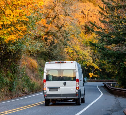 A sprinter van drives down the road among the fall leaves.