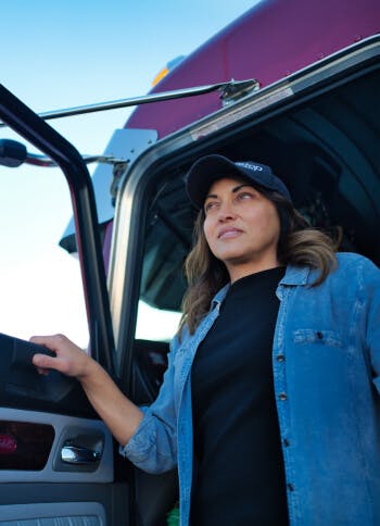 A truck driver steps out of her cab and looks out into the distance.