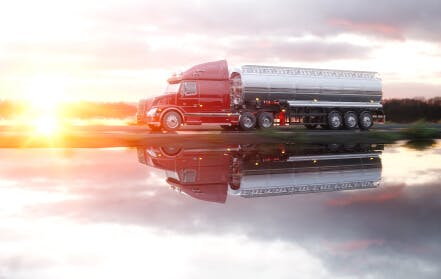 A red semi truck hauls a load along a body of water while the sun sets.
