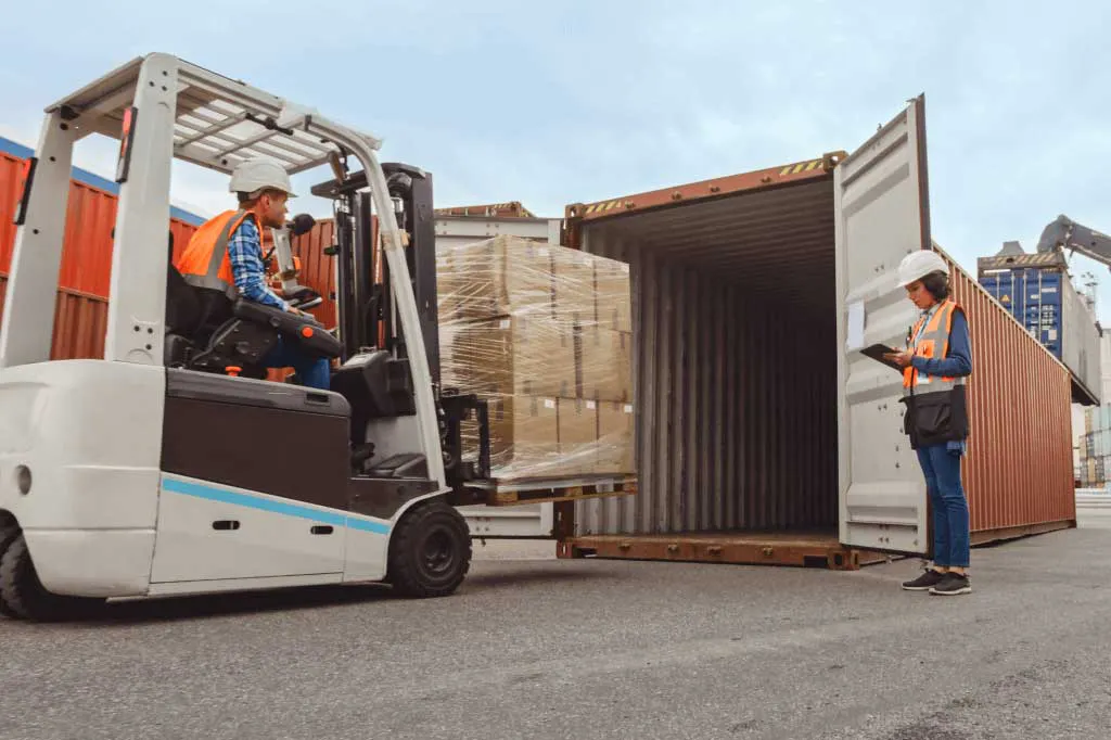 Forklift operator loading pallet into shipping container with supervisor