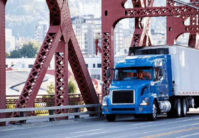 Truck and trailer crossing a bridge with a city in the background.